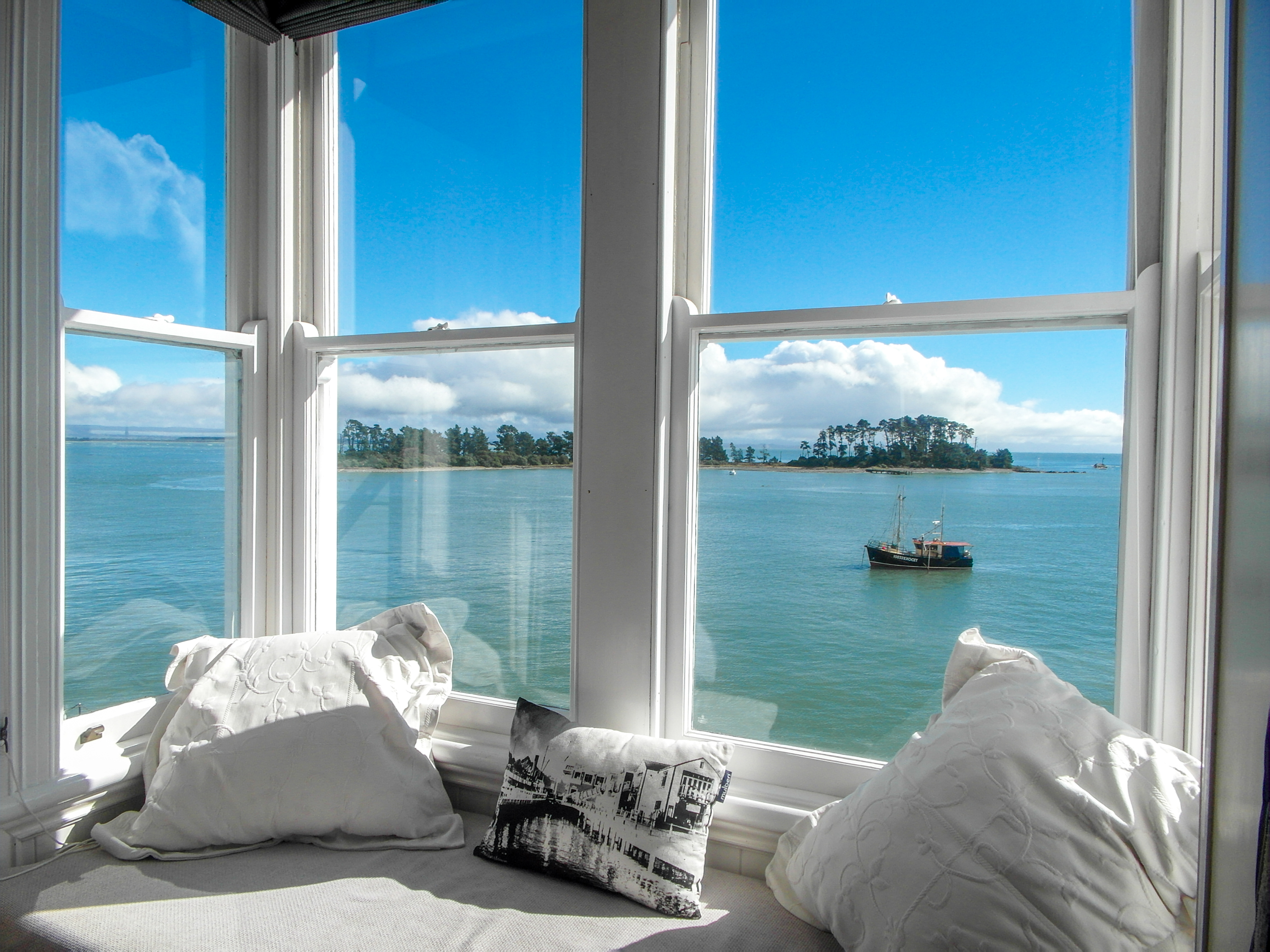 A white cushioned window seat framing blue coastal water, trees, distant land and an anchored boat.