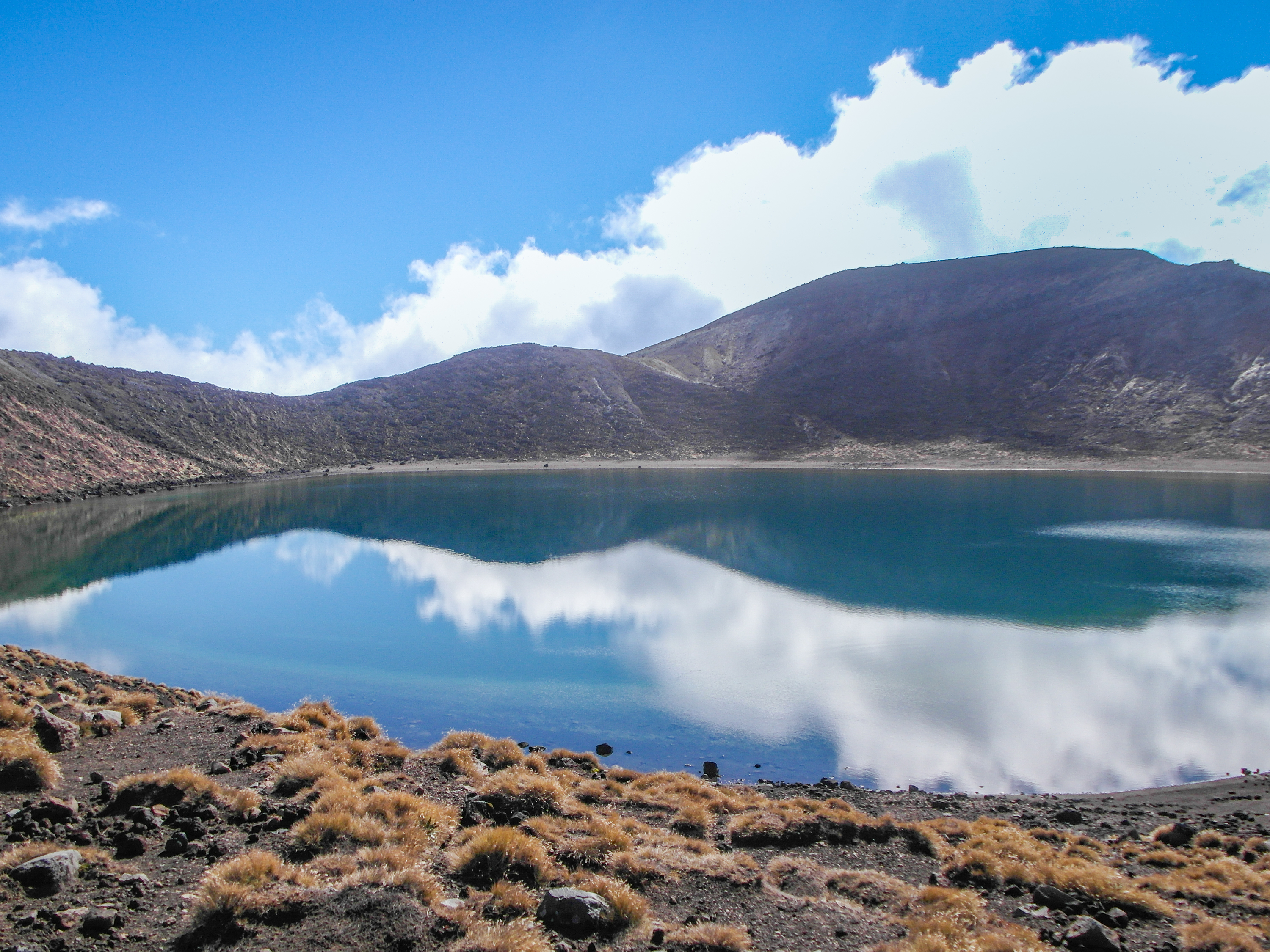 White clouds and a bare mountain rim reflected in blue lake water, with golden tussock along the near shore.