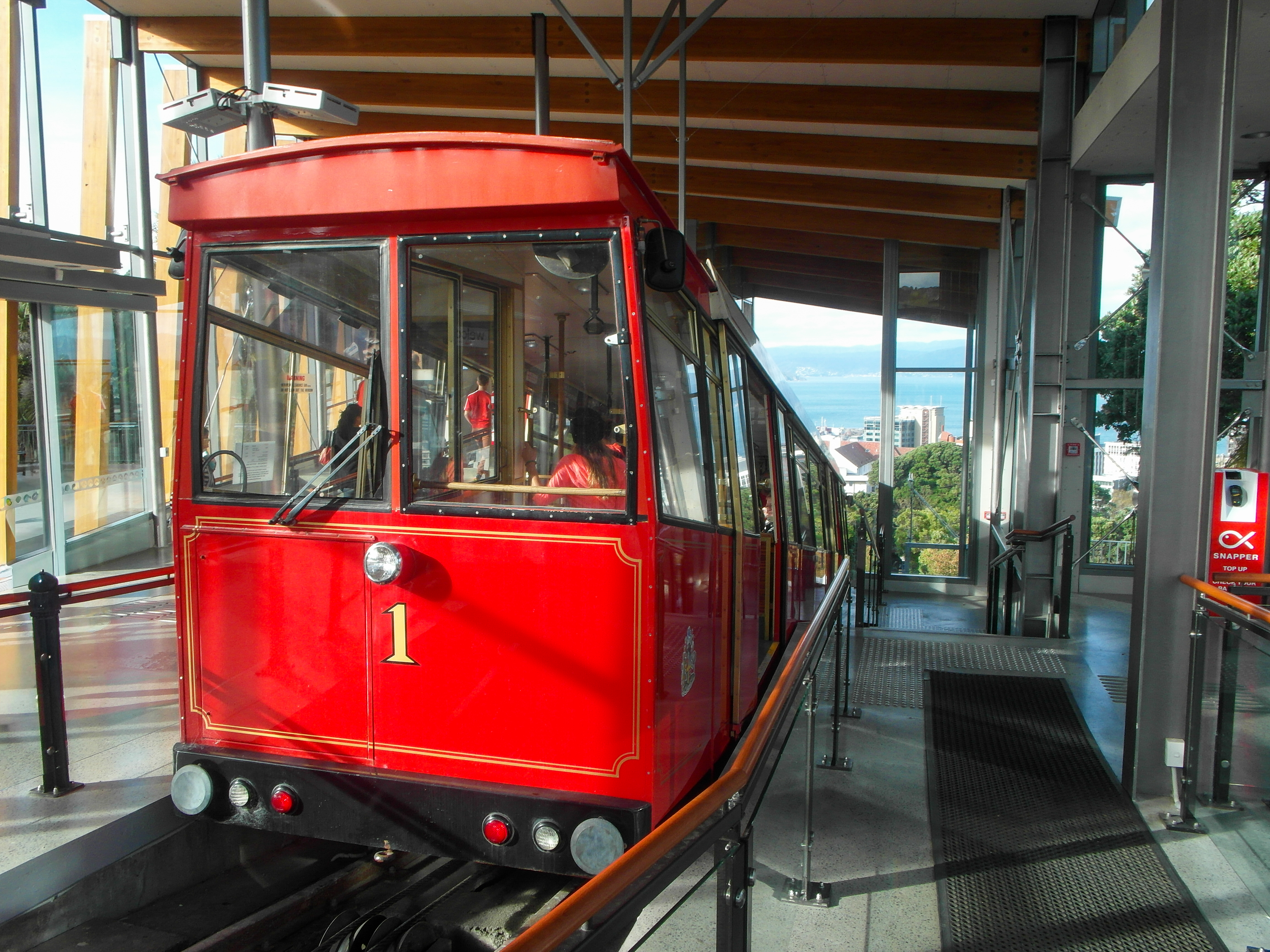 Red Wellington cable car number one at a station, with city buildings and harbour visible beyond.