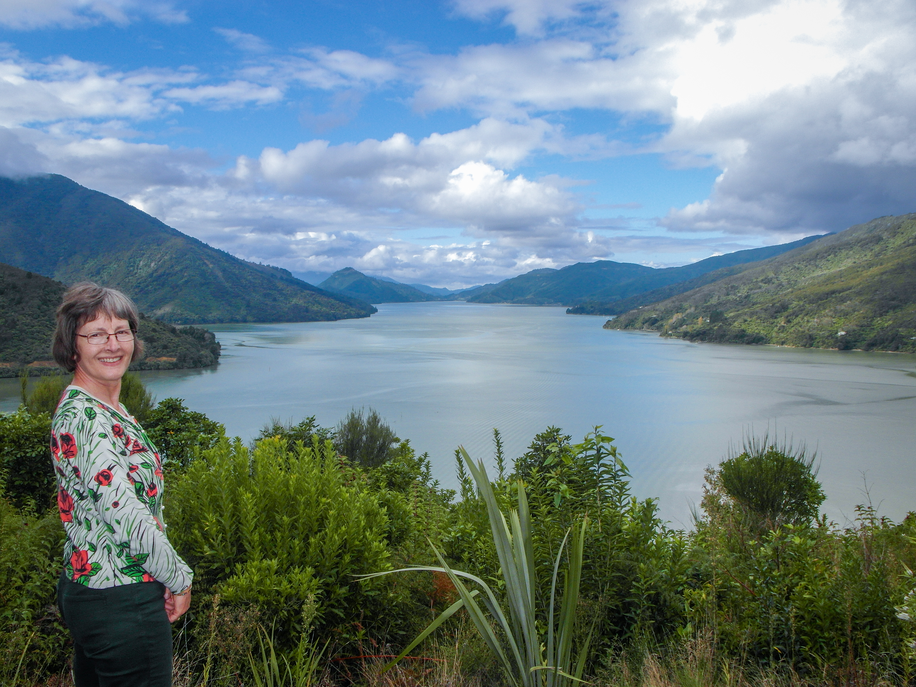 A smiling woman in a floral top standing above a broad inlet surrounded by green hills.