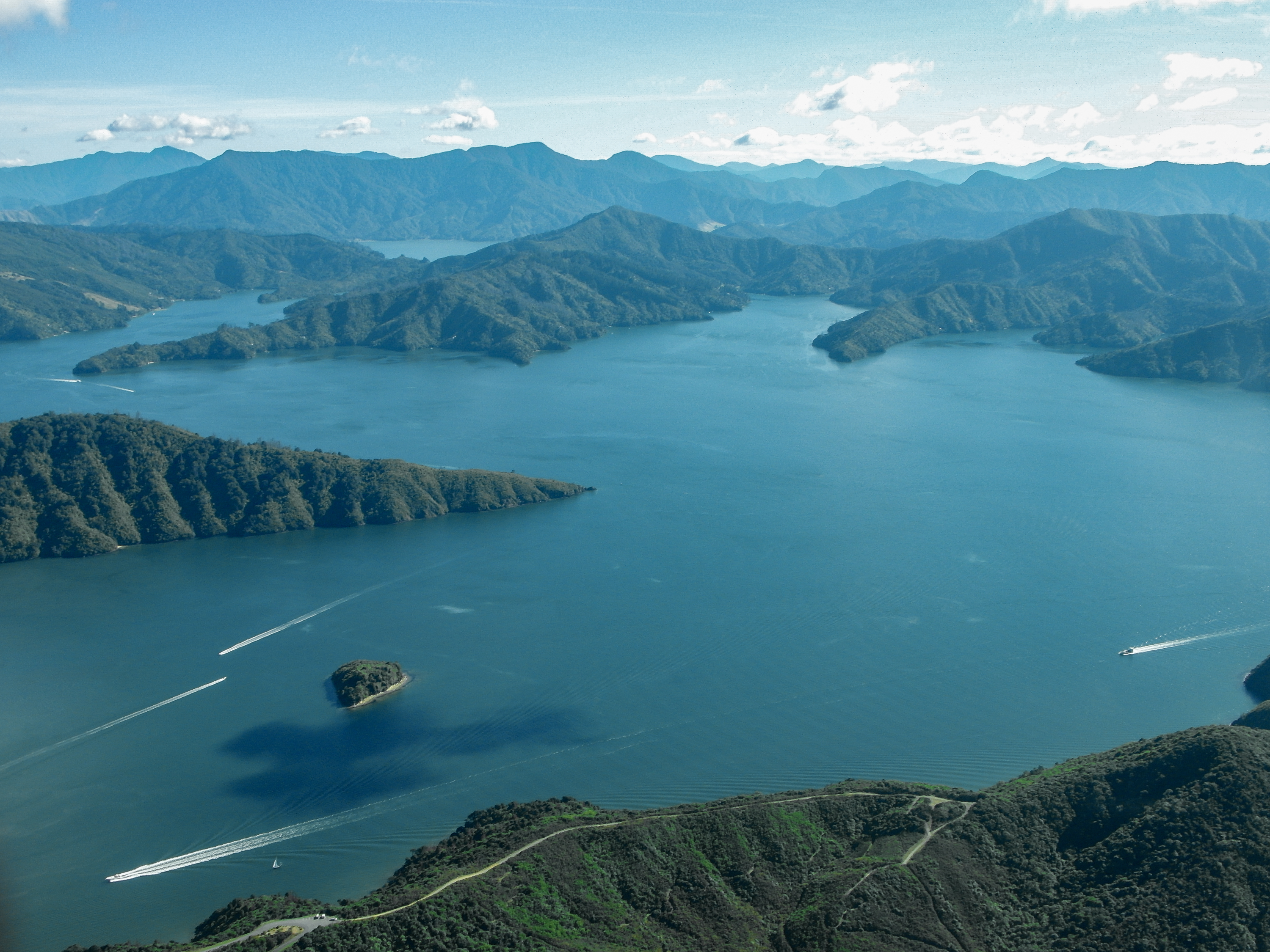 An aerial view of wooded headlands and islands surrounded by blue water with small boat wakes.
