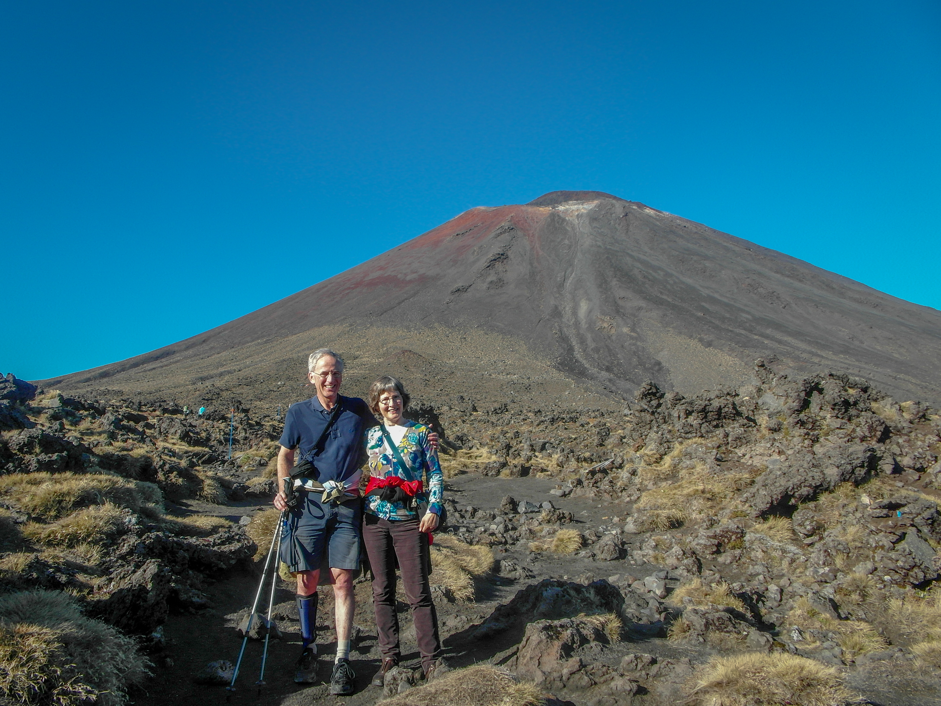 Two walkers standing among volcanic rocks and tussock below a large bare cone under a blue sky.