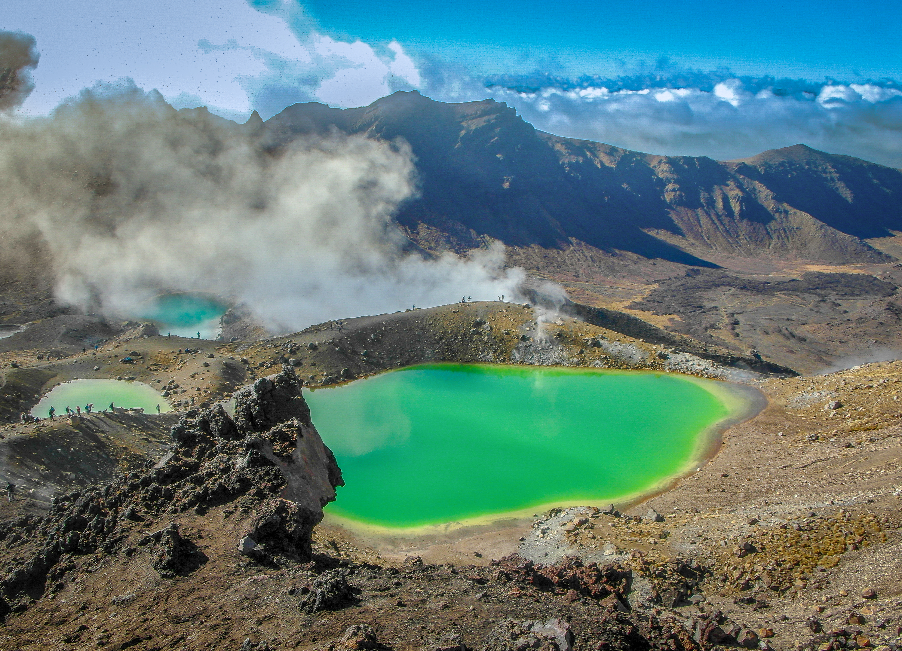 Bright green volcanic lakes surrounded by rocky ridges, with tiny walkers and drifting cloud.