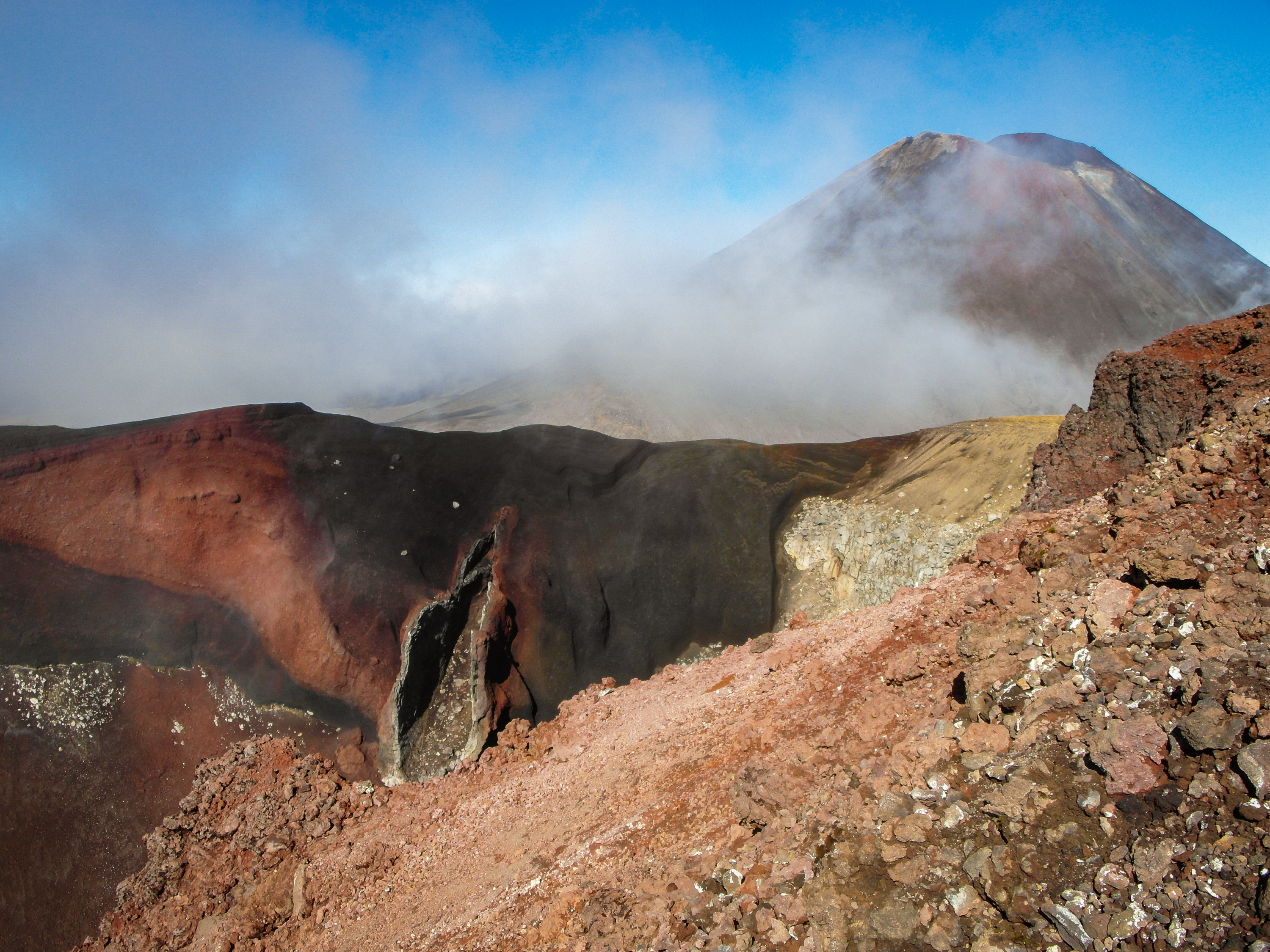 Red and black volcanic crater walls in the foreground with a mountain cone partly obscured by cloud.